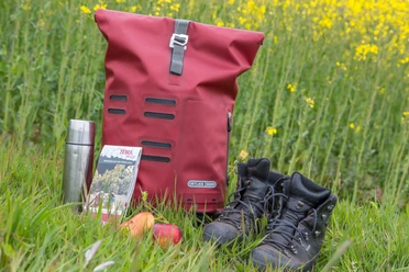 Wandeluitrusting op weide: rode rugzak, thermosfles, kaart, appel, wandelschoenen voor koolzaadveld.