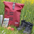 Wanderrast Wanderausrüstung auf Wiese: roter Rucksack, Thermoskanne, Karte, Apfel, Wanderschuhe vor Rapsfeld.