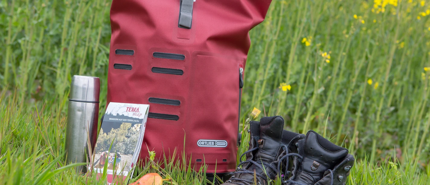 Wanderrast Hiking equipment on meadow: red rucksack, thermos flask, map, apple, hiking boots in front of rape field.