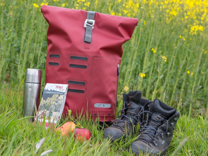 Wanderrast Wanderausrüstung auf Wiese: roter Rucksack, Thermoskanne, Karte, Apfel, Wanderschuhe vor Rapsfeld.