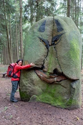 Vrouw in rode jas omhelst een groene bemoste grijnzende steen in het bos.