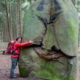 Frau in roter Jacke umarmt einen grün bemoosten grinsenden Stein im Wald.Woman in red jacket hugs a green mossy grinning stone in the forest.Kvinde i rød jakke krammer en grøn, mosgroet, grinende sten i skoven.Vrouw in rode jas omhelst een groene bemoste grijnzende steen in het bos.
