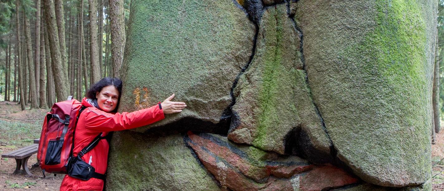 Wanderin am Süntelstein Frau in roter Jacke umarmt einen grün bemoosten grinsenden Stein im Wald.