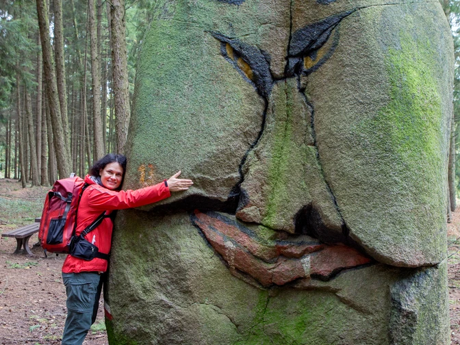 Wanderin am Süntelstein Frau in roter Jacke umarmt einen grün bemoosten grinsenden Stein im Wald.