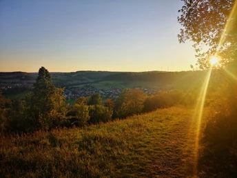 Abendstimmung am Räuschenberg bei Brenkhausen Sonnenuntergang über einer grünen Hügel Landschaft, Bäume, Wiesen und ein Dorf im Hintergrund.