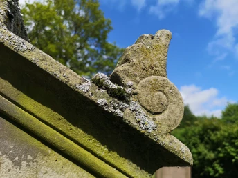 Steinerne Details auf einem Grabmonument mit Moos und Flechten, umgeben von grünen Bäumen und blauem Himmel.