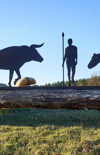 Metallskulpturen eines Mannes, eines Stiers und eines Pferdes auf einem Baumstamm in ländlicher Umgebung.
