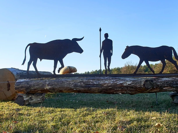 www.hoffmannfoto.de Metallskulpturen eines Mannes, eines Stiers und eines Pferdes auf einem Baumstamm in ländlicher Umgebung.