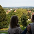 Zwei Personen genießen die Aussicht auf eine grüne Parklandschaft mit Kirchturm in der Ferne.Two people enjoy the view of a green park landscape with a church tower in the distance.To mennesker nyder udsigten over et grønt parklandskab med et kirketårn i det fjerne.Twee mensen genieten van het uitzicht op een groen parklandschap met een kerktoren in de verte.