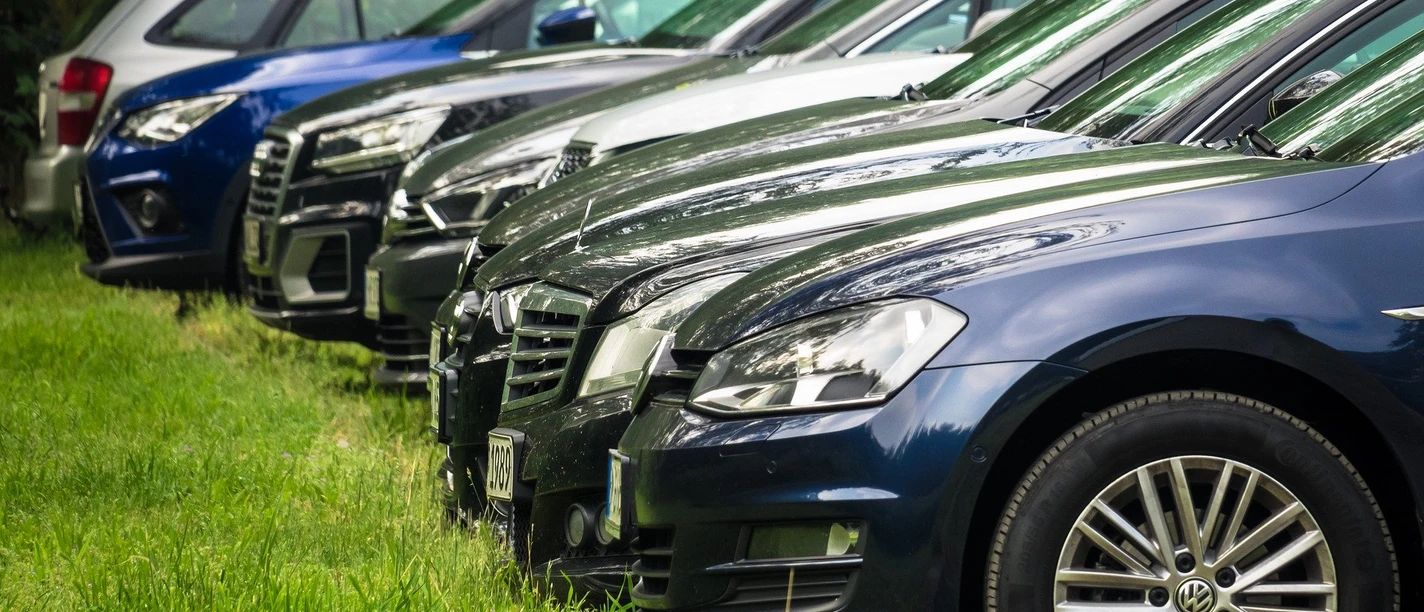 Reihe von geparkten Autos auf einer grasbewachsenen Fläche, grün umgeben. Row of parked cars on a grassy area, surrounded by greenery.