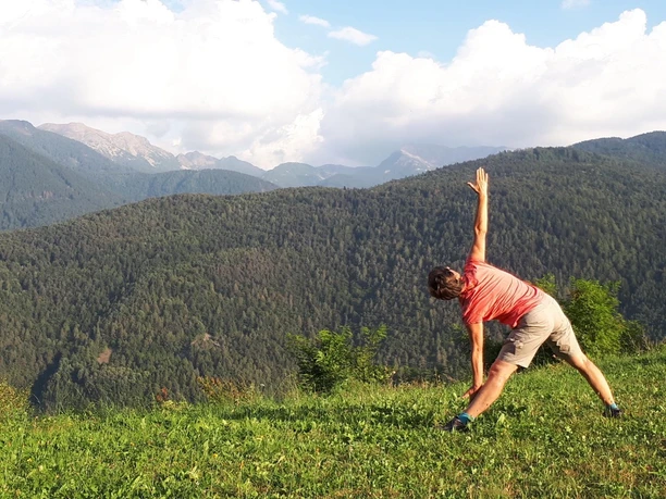 Margit Driftmeier Eine Person in Sportkleidung macht Yoga im Grünen, umgeben von bewaldeten Berghügeln.