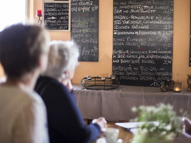 Two people sit in a restaurant and look at a wall with handwritten menu boards. The atmosphere is cozy.