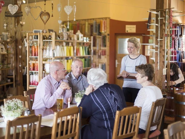 A group of elderly people sit at a wooden table in a cozy store with colorful candles in the background while a woman serves them.