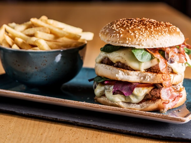 Hamburger met friet Dubbele cheeseburger met bacon, salade en kaas op een bord, naast een kom friet, geserveerd op een houten tafel.