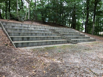 Steingestufte Sitzreihen einer Freilichtbühne umgeben von dicht bepflanzten Bäumen im Grünen.