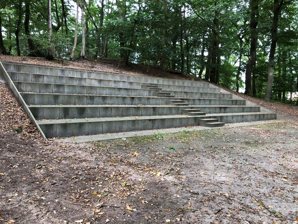 Steingestufte Sitzreihen einer Freilichtbühne umgeben von dicht bepflanzten Bäumen im Grünen.