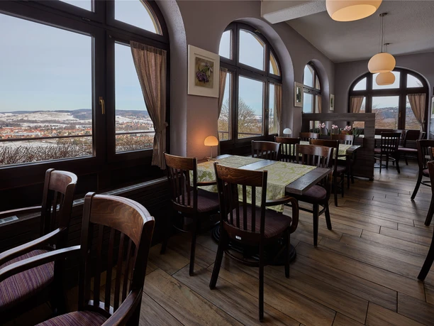 Cozy café with wooden furniture and large windows offering a view of a snowy landscape; warm lighting and decorative plants.