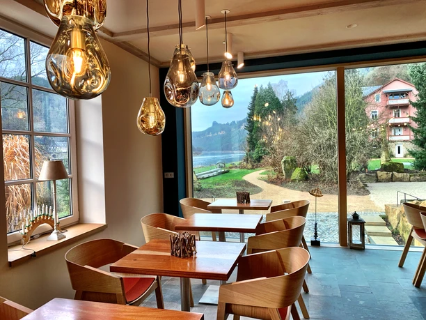 Cozy café with wooden furniture and large windows offering a view of a snowy landscape; warm lighting and decorative plants.