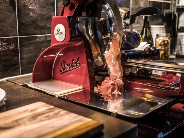 Red Berkel slicer cutting thin slices of ham; tiled wall and kitchen utensils in the background.