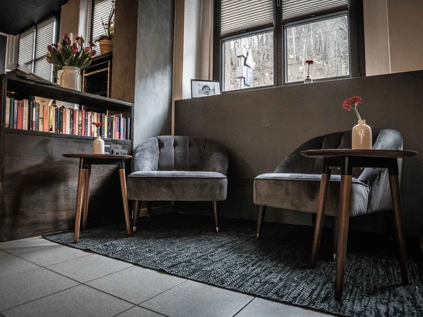 Comfortable seating area with two gray armchairs, a wooden table with a red flower in a vase, bookshelf and window in the background.