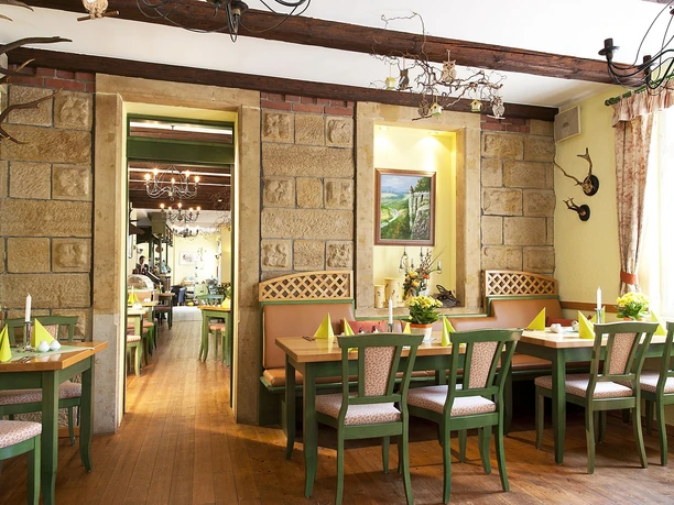 Gezellig restaurant met houten meubels en groene stoelen, versierd met kaarsen en bloemen; stenen muren en een schilderij op de achtergrond.