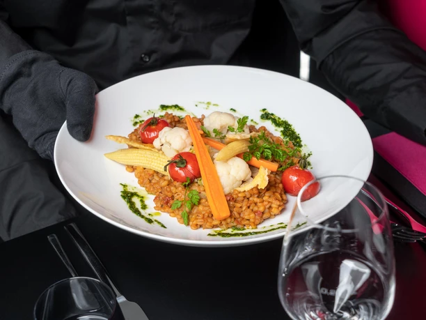 A plate of barley risotto, decorated with baby corn, carrots, cauliflower, cherry tomatoes and herbs, is held by a person in black clothing.