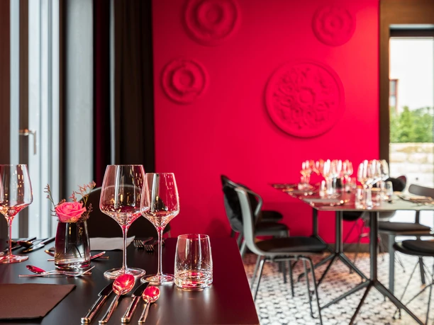 Elegant restaurant with red walls, laid tables, wine glasses and a pink flower in a vase. Modern, stylish atmosphere.