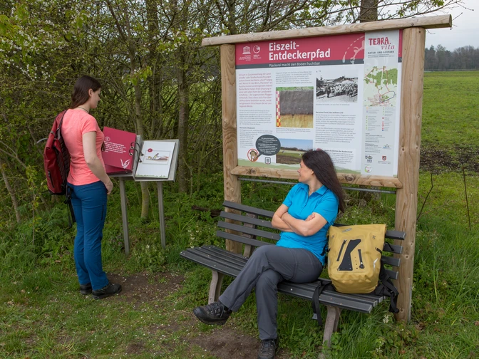TERRA.track Eiszeit-Entdeckerpfad Zwei Frauen erkunden einen Lehrpfad mit Infotafeln über die Eiszeit in einer ländlichen Umgebung.Two women explore an educational trail with information boards about the Ice Age in a rural setting.To kvinder udforsker en undervisningssti med informationstavler om istiden i landlige omgivelser.Twee vrouwen verkennen een educatief pad met informatieborden over de ijstijd in een landelijke omgeving.