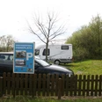 Stellplatz mit Schild für Wohnmobile und Pkw, umgeben von Bäumen und Wiese mit Löwenzahn.