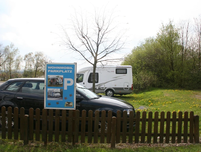 Stellplatz mit Schild für Wohnmobile und Pkw, umgeben von Bäumen und Wiese mit Löwenzahn.