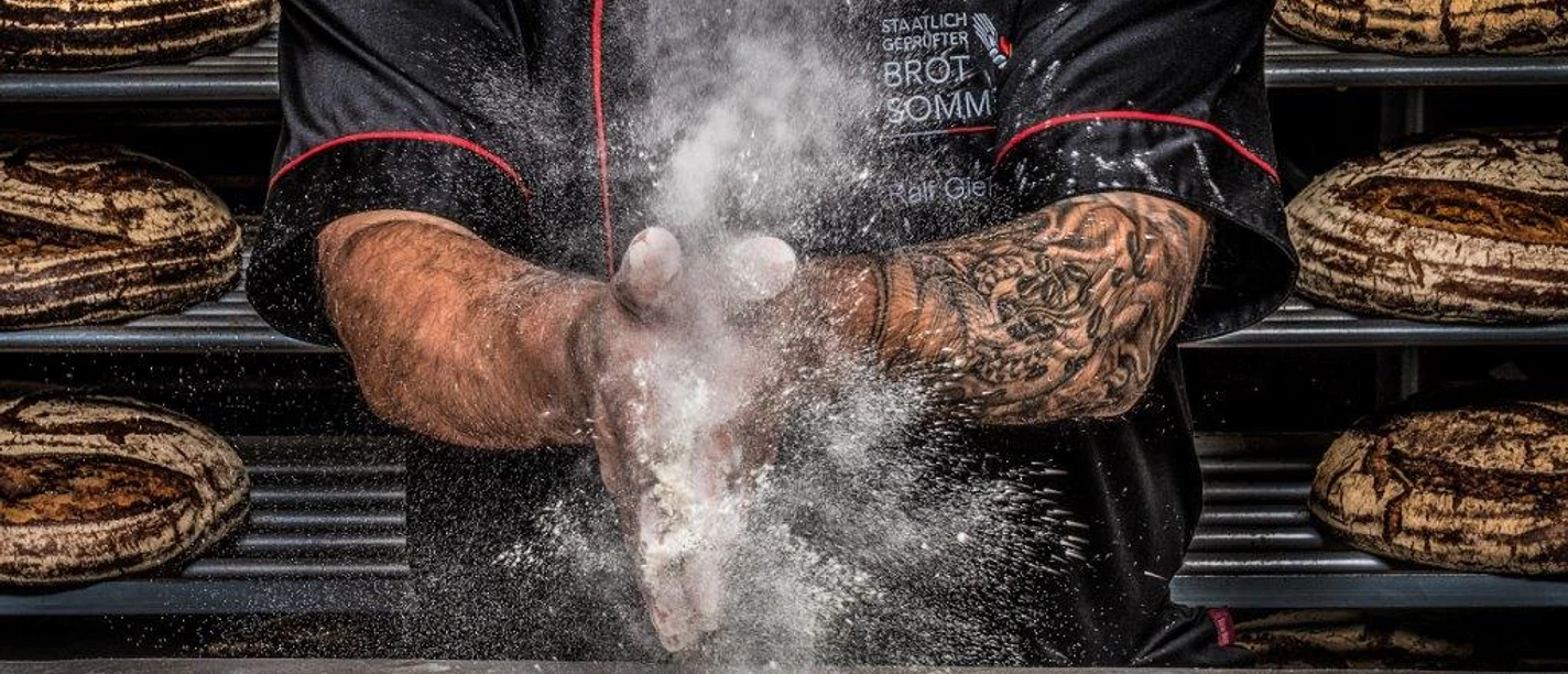 Bäckerei Gießelmann Ein Bäcker in schwarzer Kleidung wirbelt Mehl in die Luft, umgeben von frisch gebackenem Brot.