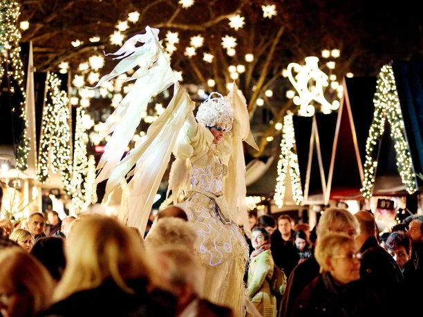 Markt der Engel in Köln Stimmungsvoll beleuchteter Weihnachtsmarkt „Markt der Engel“ auf dem Kölner Neumarkt. Im Zentrum hat eine majestätische Engelsfigur mit prächtigen Flügeln eine erhöhte Position eingenommen, umringt von einem lebhaften Meer aus Menschen und funkelnden Lichtern. Die Szenerie vermittelt eine festliche und magische Atmosphäre, die Besucher verzaubert.