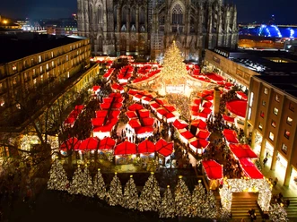 Christmas market at Cologne Cathedral A festively illuminated Christmas market with numerous red sales huts under the majestic Cologne Cathedral at night. The market is crowded, the atmosphere is lively and cheerful. A large, brightly decorated fir tree dominates the scene.