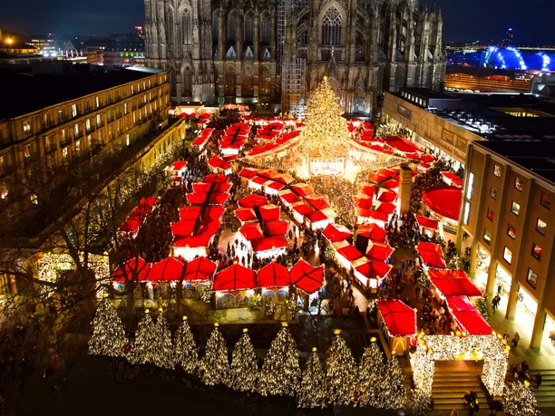 Christmas market at Cologne Cathedral A festively illuminated Christmas market with numerous red sales huts under the majestic Cologne Cathedral at night. The market is crowded, the atmosphere is lively and cheerful. A large, brightly decorated fir tree dominates the scene.