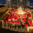 Weihnachtsmarkt am Kölner Dom Ein festlich beleuchteter Weihnachtsmarkt mit zahlreichen roten Verkaufshütten unter dem majestätischen Kölner Dom bei Nacht. Der Markt ist dicht besucht, die Atmosphäre ist lebendig und fröhlich. Eine große, strahlend geschmückte Tanne dominiert die Szene.A festively illuminated Christmas market with numerous red sales huts under the majestic Cologne Cathedral at night. The market is crowded, the atmosphere is lively and cheerful. A large, brightly decorated fir tree dominates the scene.