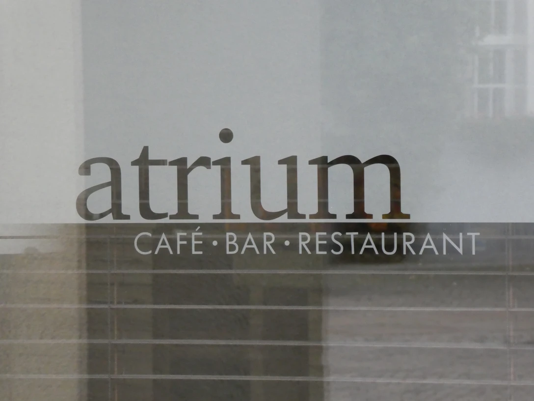 Schriftzug "Atrium Café Bar Restaurant" auf einer Glasoberfläche, dahinter verschwommene Gebäudestrukturen.Lettering "Atrium Café Bar Restaurant" on a glass surface, behind it blurred building structures.Bogstaverne "Atrium Café Bar Restaurant" på en glasoverflade, bagved slørede bygningsstrukturer.Belettering "Atrium Café Bar Restaurant" op een glazen oppervlak, daarachter vage gebouwstructuren.
