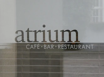 Atrium Schriftzug "Atrium Café Bar Restaurant" auf einer Glasoberfläche, dahinter verschwommene Gebäudestrukturen.