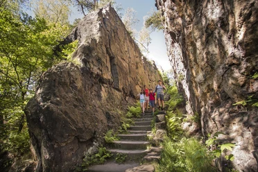 Zittauer Gebirge_Katja Fouad Vollmer (16).JPG Drei Wanderer auf einem Pfad zwischen hohen Felswänden und grüner Vegetation im Zittauer Gebirge.