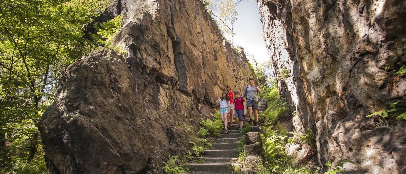 Zittauer Gebirge_Katja Fouad Vollmer (16).JPG Drei Wanderer auf einem Pfad zwischen hohen Felswänden und grüner Vegetation im Zittauer Gebirge.