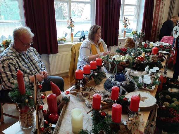 Weihnachtsmarkt Tange.jpg Menschen verkaufen Adventsgestecke mit roten Kerzen und Tannenzweigen auf einem Weihnachtstisch.