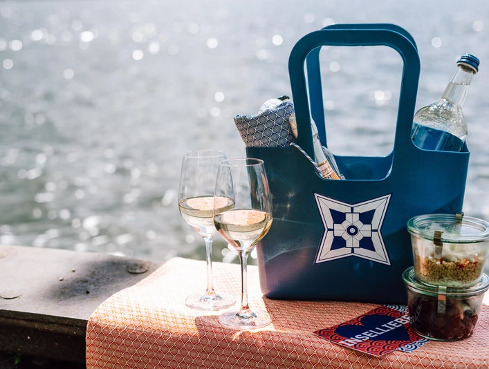 Picknick Zwei Gläser Weißwein, ein gefüllter Picknickkorb und Plastikbehälter auf einem Tuch am Seeufer.Two glasses of white wine, a filled picnic basket and plastic containers on a cloth on the lakeshore.To glas hvidvin, en fyldt picnickurv og plastikbeholdere på et klæde ved søbredden.Twee glazen witte wijn, een gevulde picknickmand en plastic bakjes op een doek aan de oever van het meer.