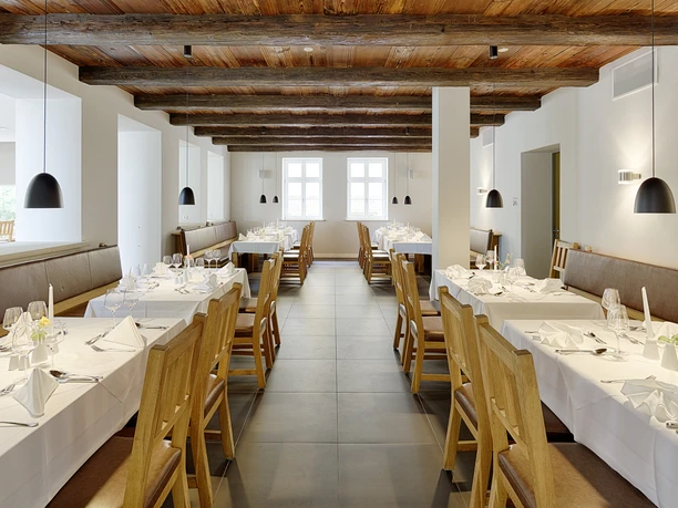 Světlá restaurace s dřevěným trámovým stropem, stoly s bílými ubrusy a dřevěnými židlemi, osvětlená černými závěsnými lampami.