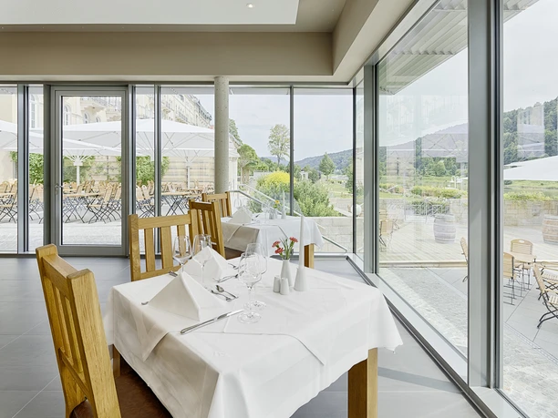 Elegantní restaurace s dřevěným nábytkem a bíle prostřenými stoly má velká okna s výhledem na terasu se slunečníky a zelenou krajinu.