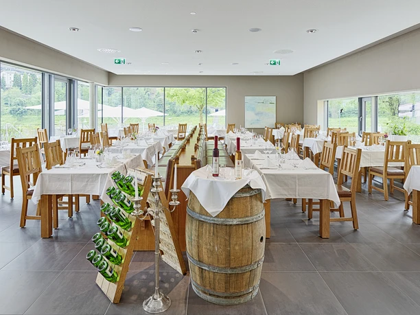 Světlá restaurace s dřevěným nábytkem, bíle prostřenými stoly, sudem na víno a stojanem na lahve; z velkých oken je výhled do zeleně.