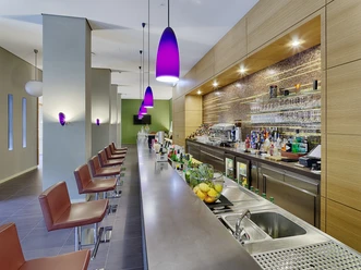 Hotel Elbresidenz Rio Xingu Bar Moderne Bar mit langer Theke, lila Hängelampen, roten Barhockern und einer gut bestückten Rückwand mit Flaschen und Gläsern.