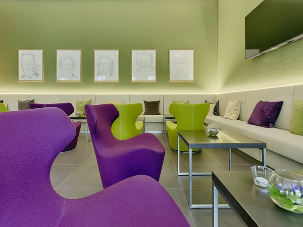 Modern waiting room with purple and green armchairs, beige sofas, colorful cushions and drawings on the green wall. Calm, inviting atmosphere.