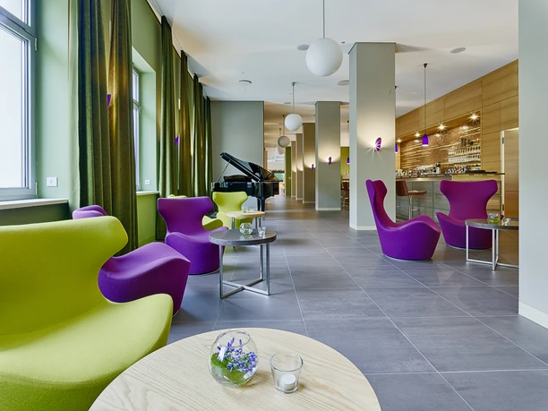Modern lounge with purple and green armchairs, round tables, a grand piano and a bar in the background. Bright, relaxed atmosphere.