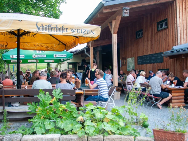 Een biertuin met gasten aan houten tafels onder parasols, omgeven door groene natuur.