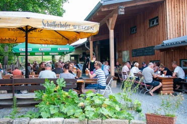 Ottendorfer Hütte Biergarten Ein Biergarten mit Gästen an Holztischen unter Sonnenschirmen, umgeben von grüner Natur.