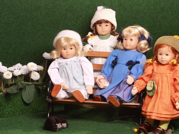 Puppenstube - Freunde Vier kunstvoll gekleidete Puppen sitzen auf einer Bank in einer blühenden Gartenkulisse.